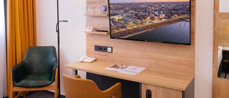 Habitación de hotel con un escritorio de madera, un televisor de pantalla plana mostrando un paisaje urbano, una silla verde y una silla marrón.