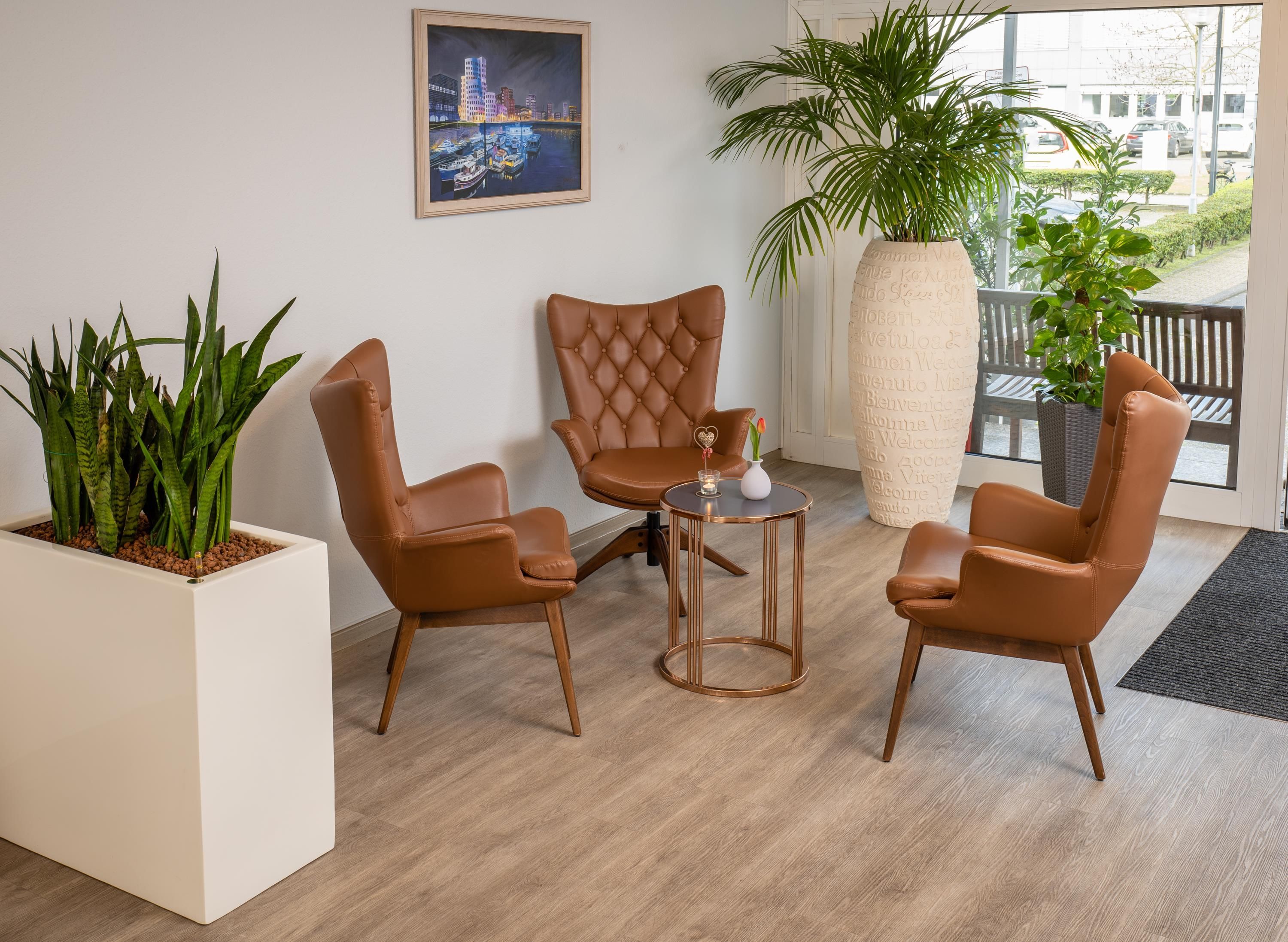 A room with three brown leather chairs, a small round table, a potted plant, and a framed cityscape painting on the wall.