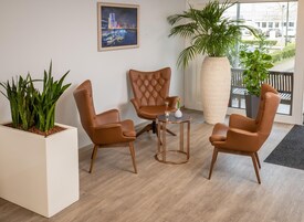 Una habitación con tres sillones de cuero marrón, una mesita redonda, una planta en maceta y un cuadro con una vista urbana en la pared.
