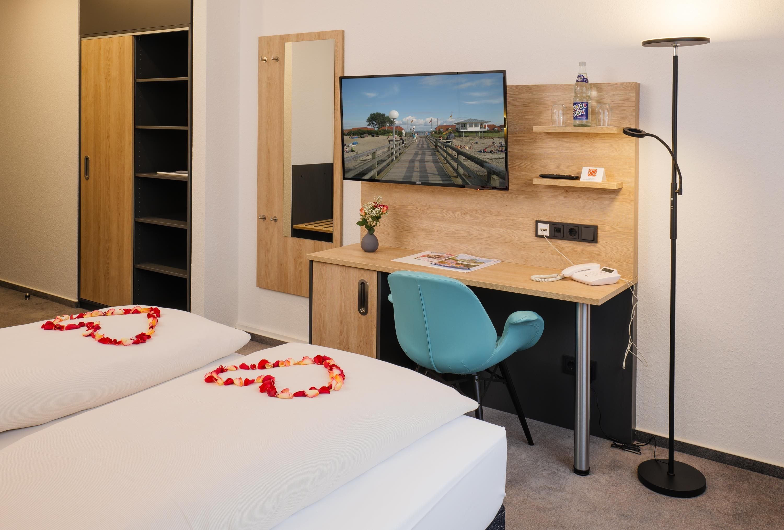 A hotel room with a bed, a desk with a computer monitor, a chair, a telephone, and a bedside table with a bottle and glasses.