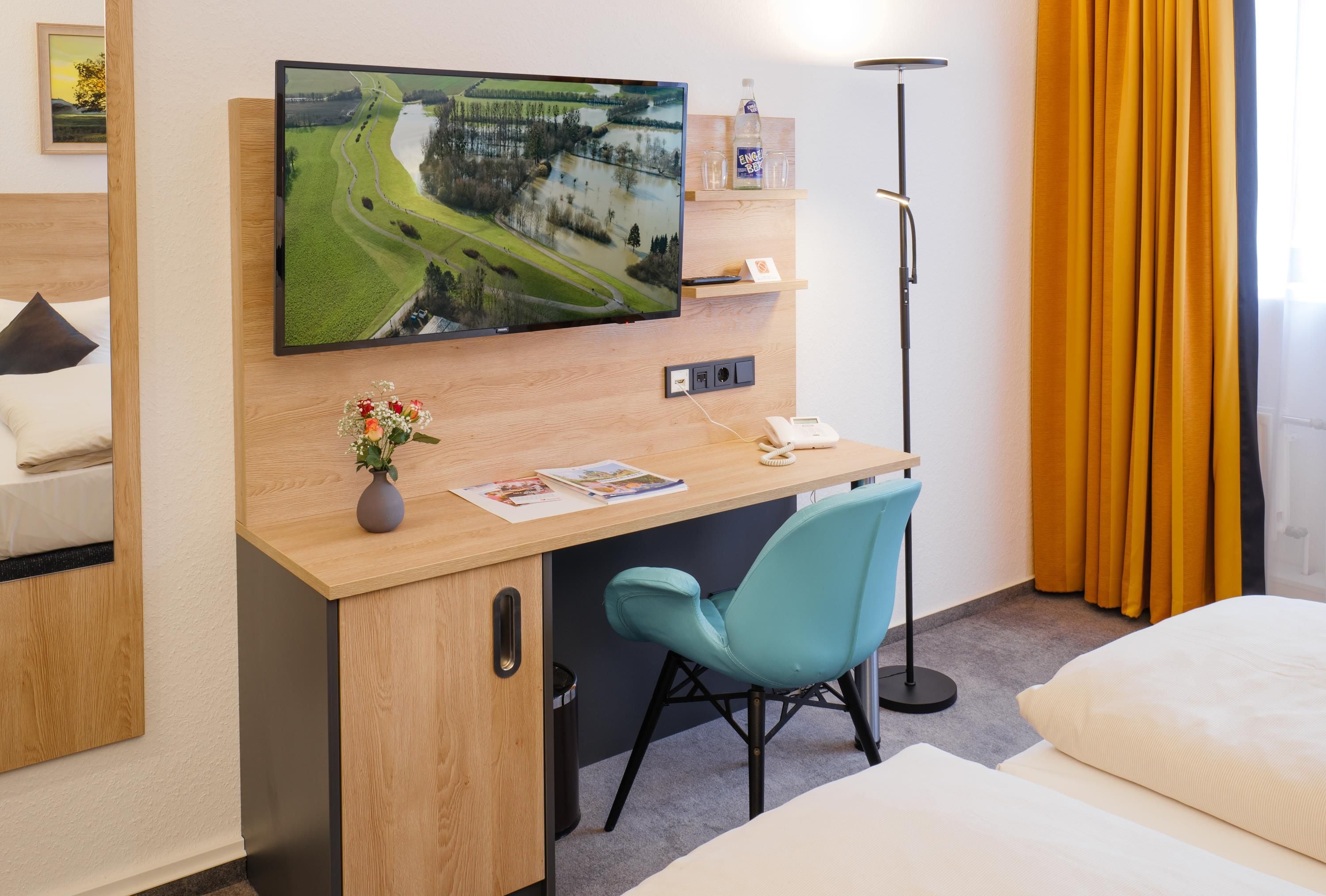 A hotel room with a wooden desk, a blue chair, a bed, a TV mounted on the wall, and a floor lamp.