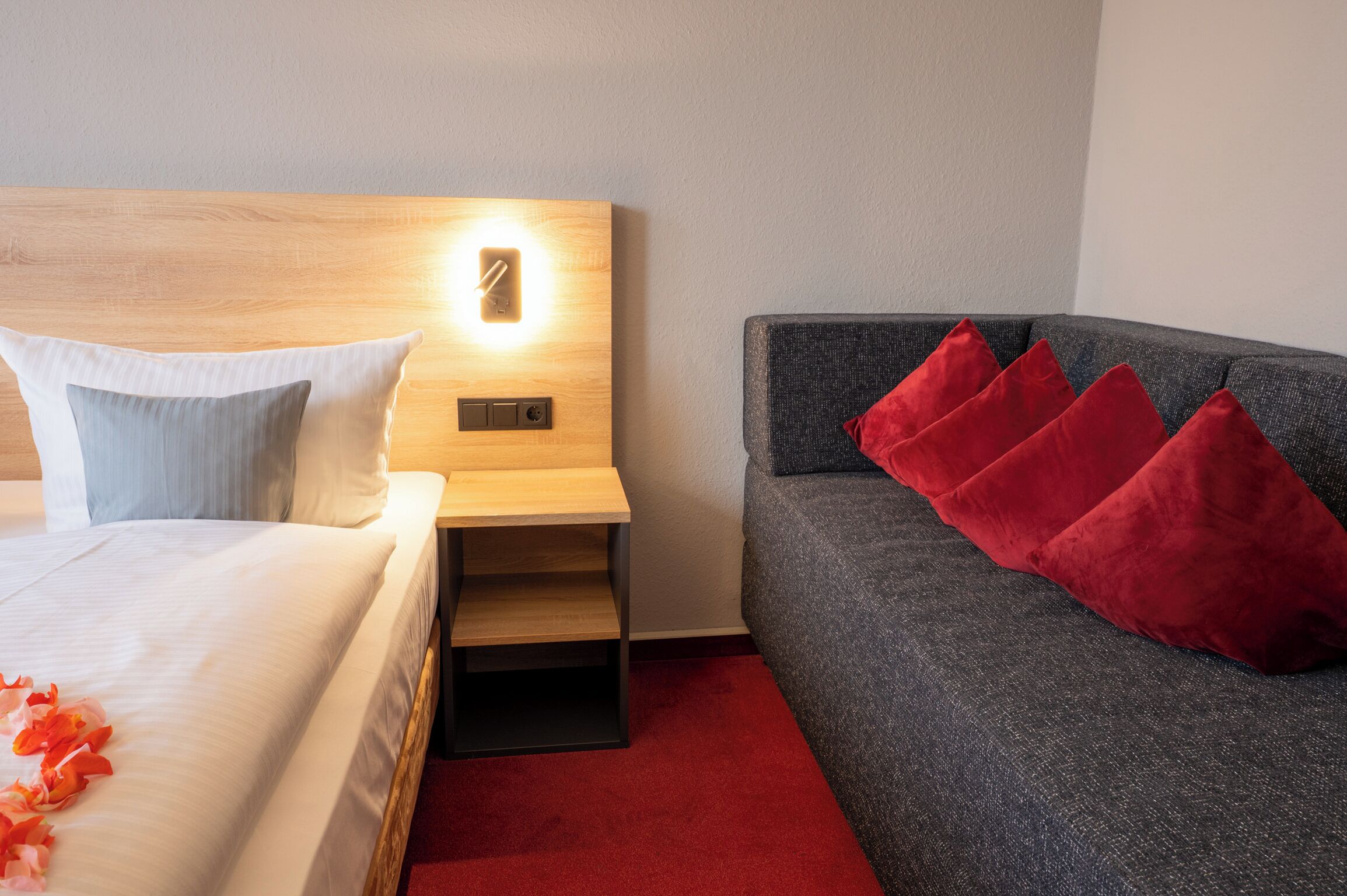 Una habitación de hotel con cama, mesita de noche y un sofá gris con cojines rojos.