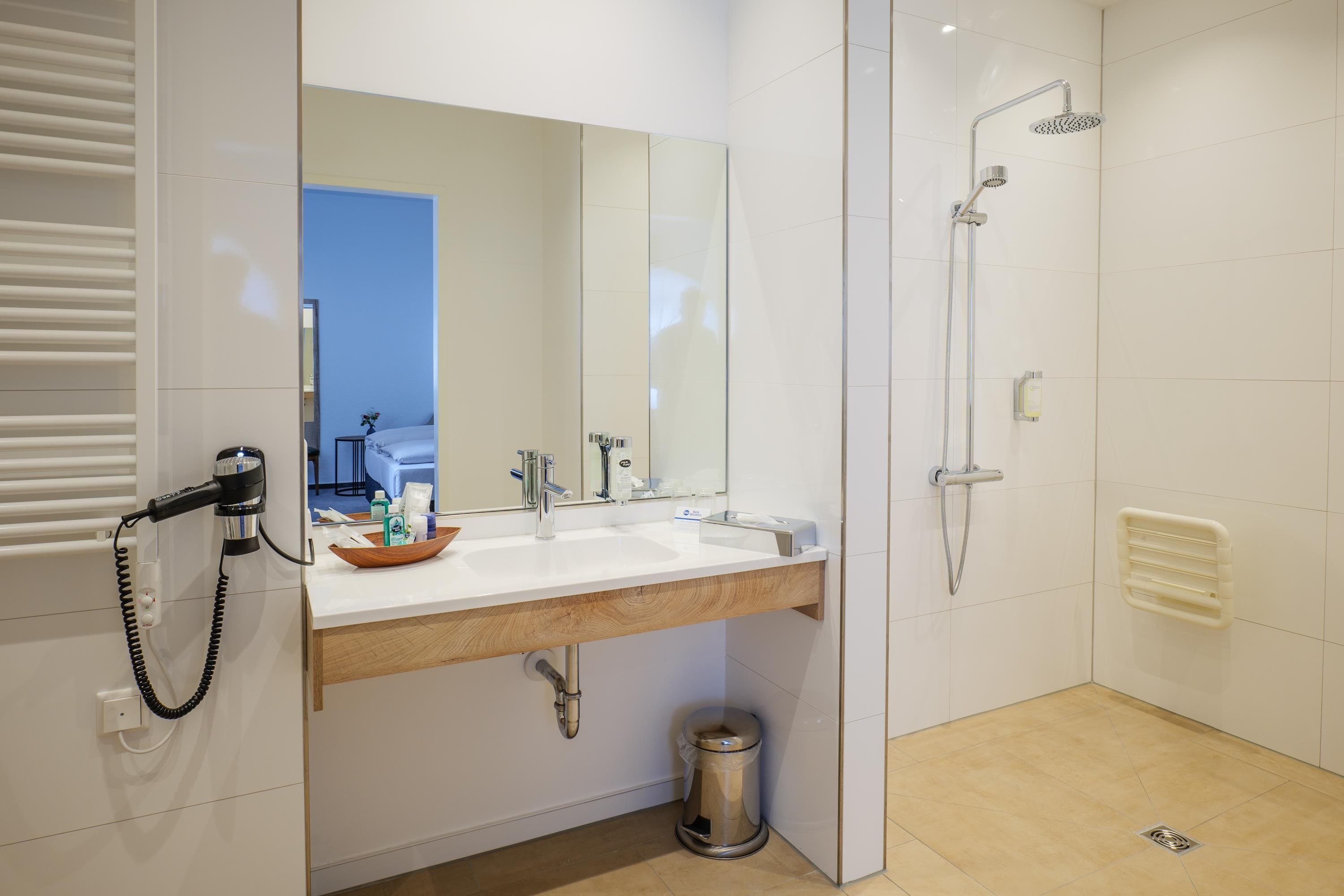 Ein modernes Badezimmer mit einem weißen Waschtisch, einem großen Spiegel, einer Dusche inklusive Handbrause und einem Mülleimer.