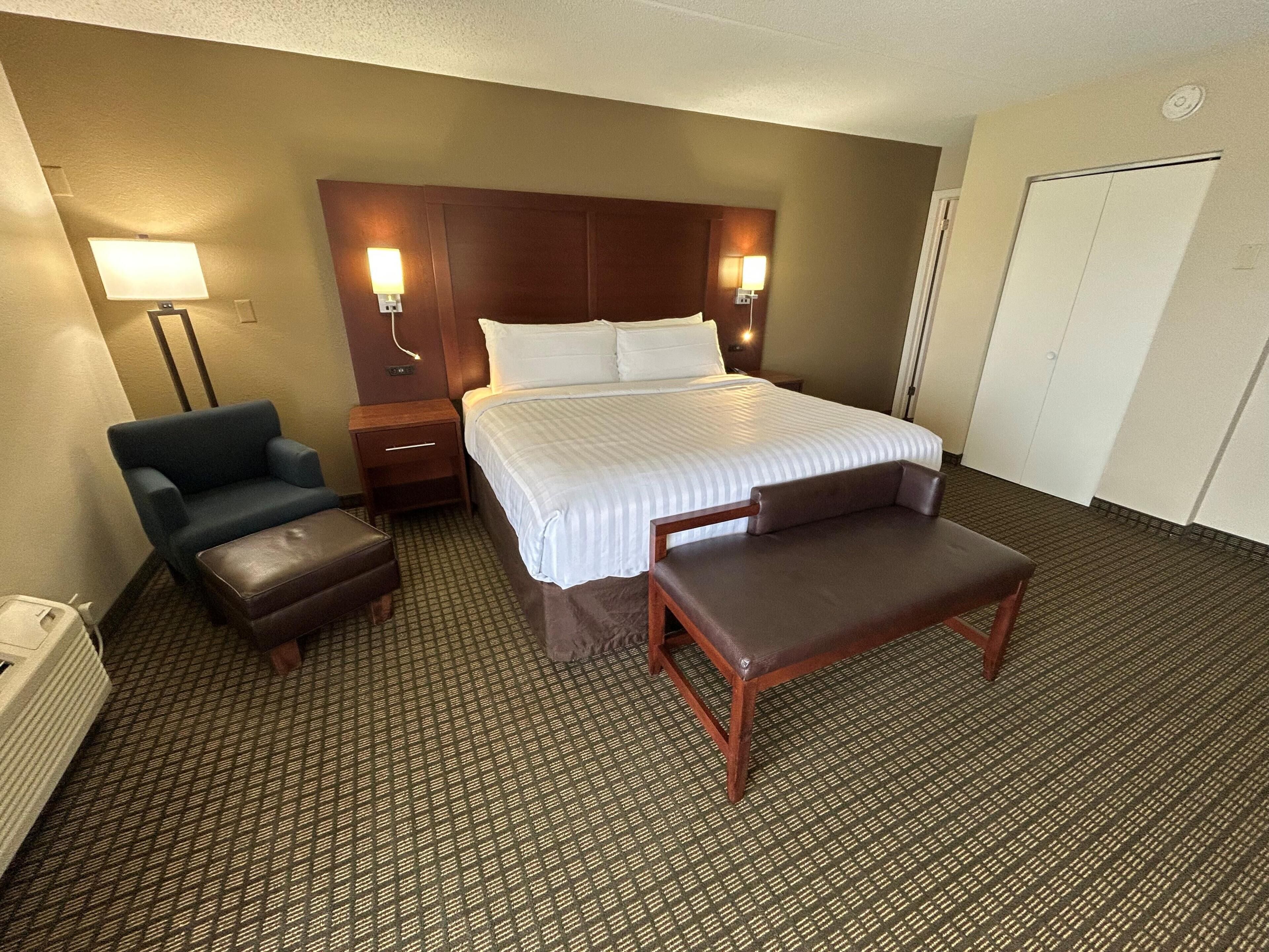 Una habitación de hotel con cama, una silla, un banco, una mesita de noche y una lámpara.