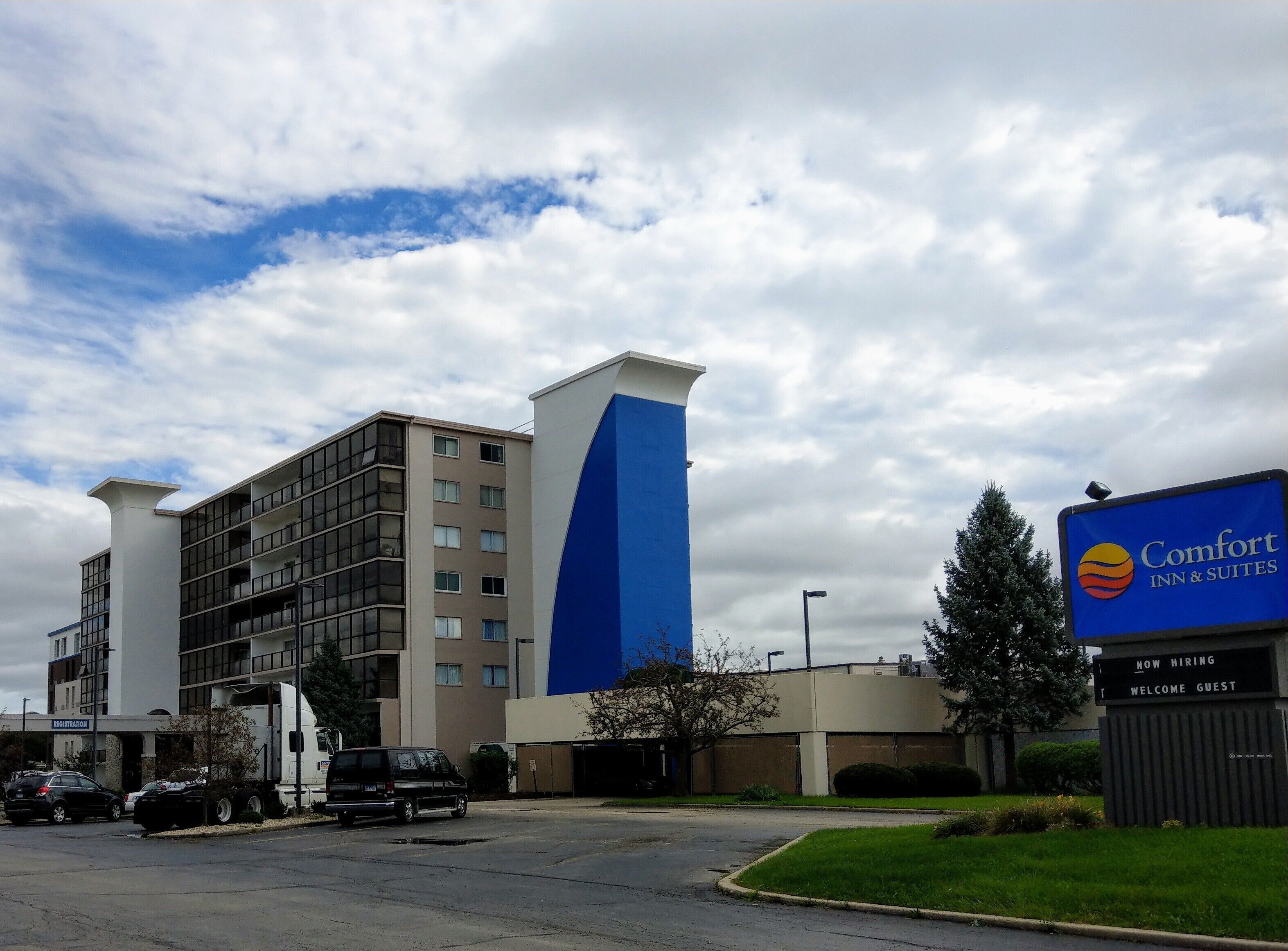 Een Comfort Inn & Suites hotel met een modern design, gekenmerkt door een opvallend blauw-beige kleurenschema, een parkeerplaats met meerdere voertuigen, en een heldere lucht met wolken.