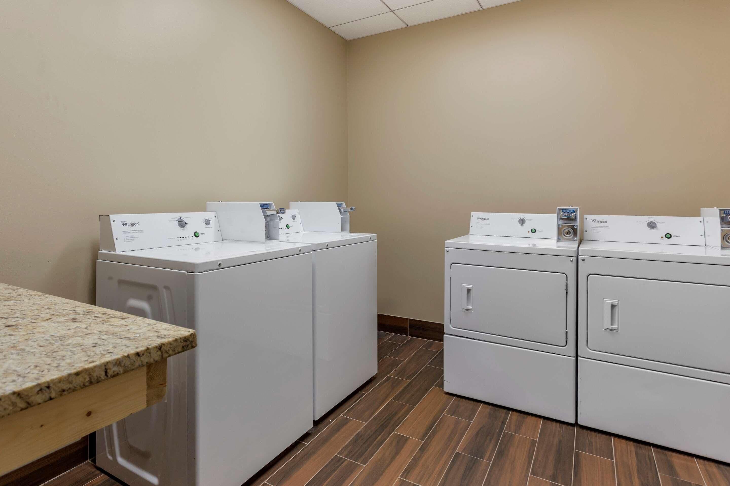 Une buanderie équipée de deux lave-linge et deux sèche-linge Whirlpool, d’un plan de travail en granit et d’un revêtement de sol en bois.