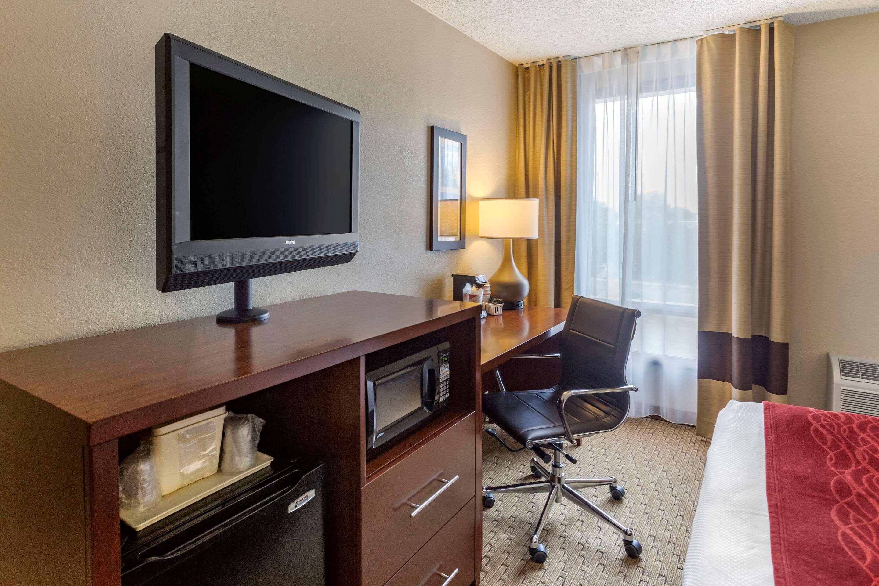 A hotel room with a flat-screen TV, a desk with a lamp, a chair, a bed with a red blanket, and a window with curtains.