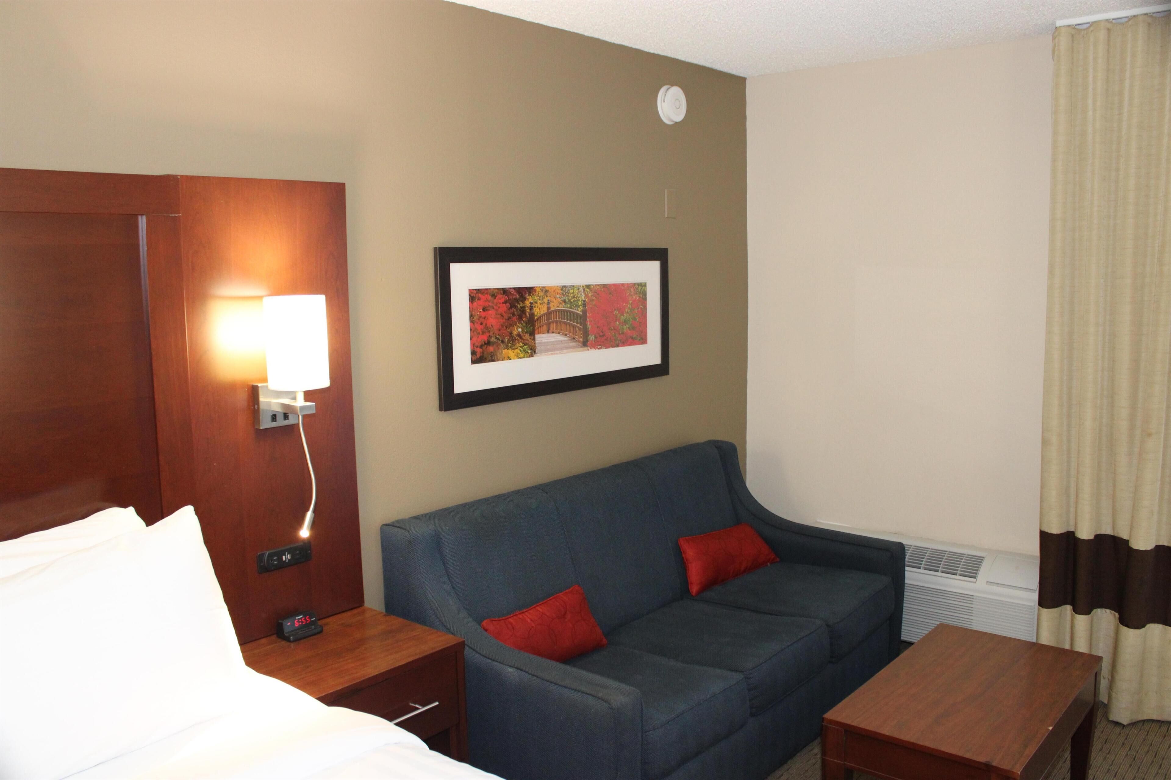 A hotel room with a bed, a blue sofa with red cushions, a small table, a framed picture on the wall, a nightstand with a lamp, and a wall-mounted air conditioning unit.