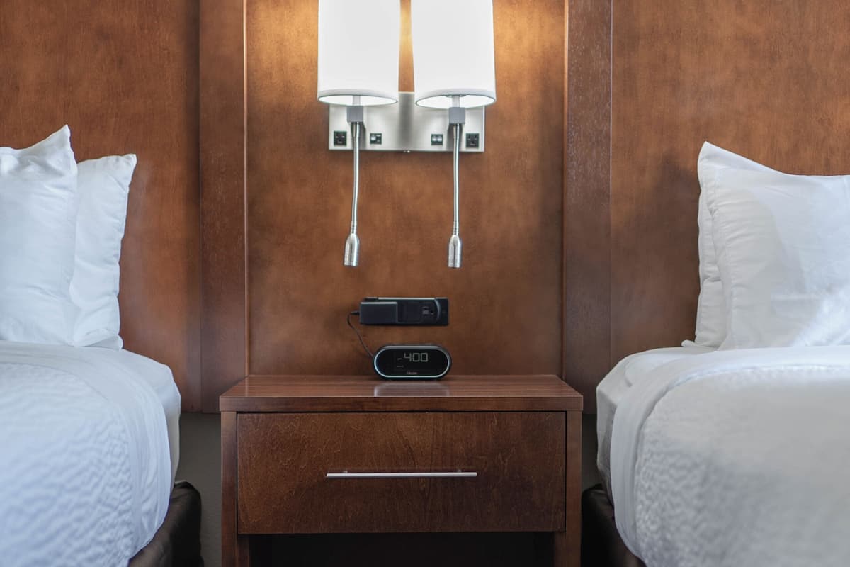 A hotel room with two beds, a wooden nightstand with a lamp, and a small drawer.