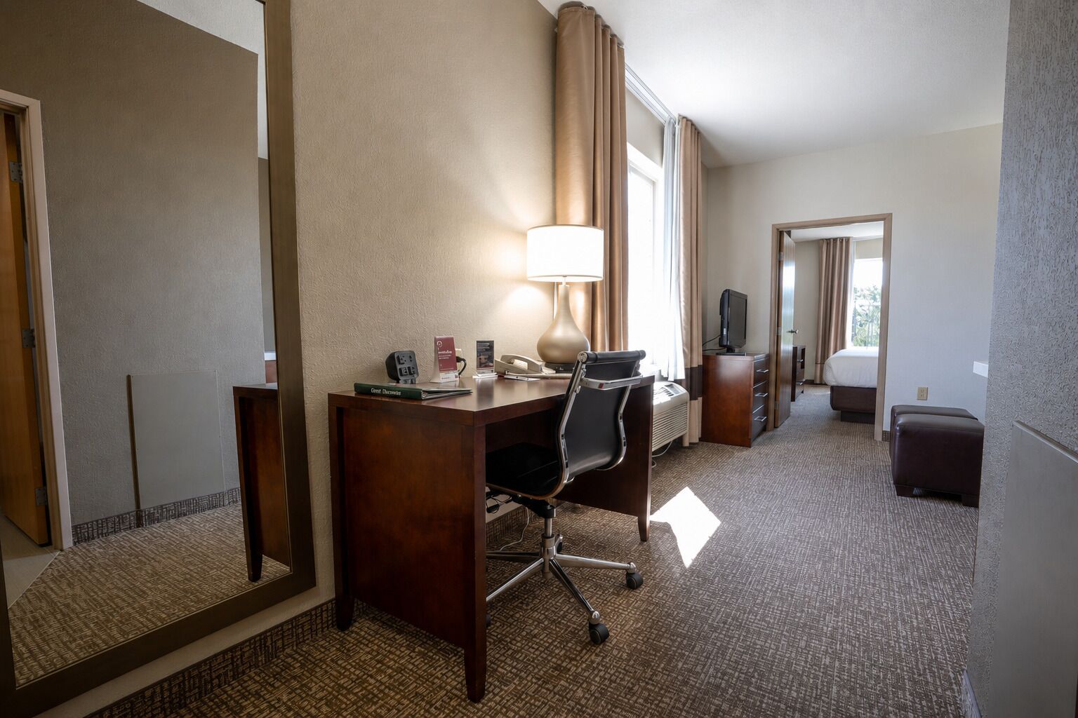 A hotel room with a desk, chair, lamp, television, and a large mirror.