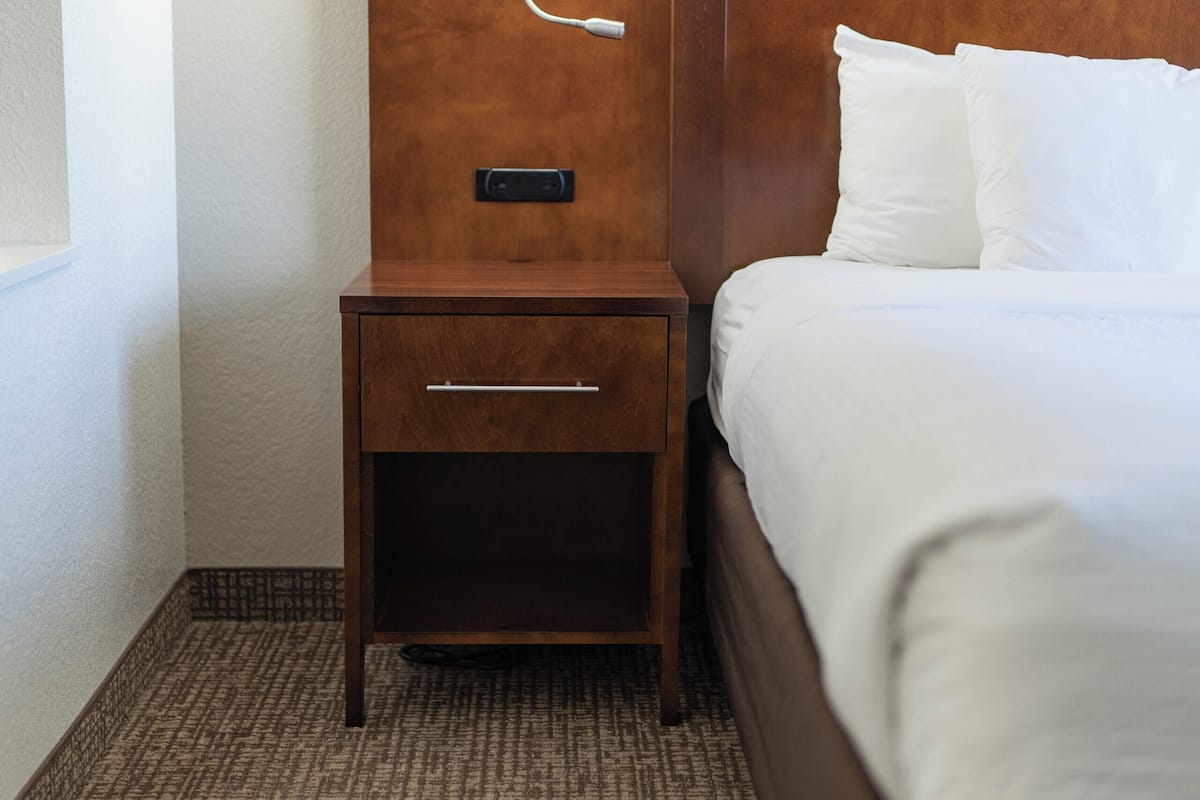 A hotel room with a wooden headboard, a bedside table with a lamp, a drawer, and a bed with white bedding.