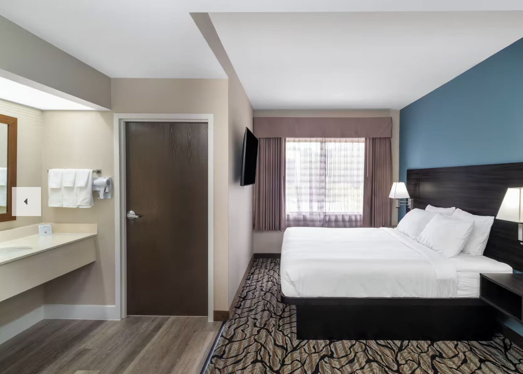 A hotel room with a bed, a TV, a bathroom with a mirror and towel rack, and a window with curtains.