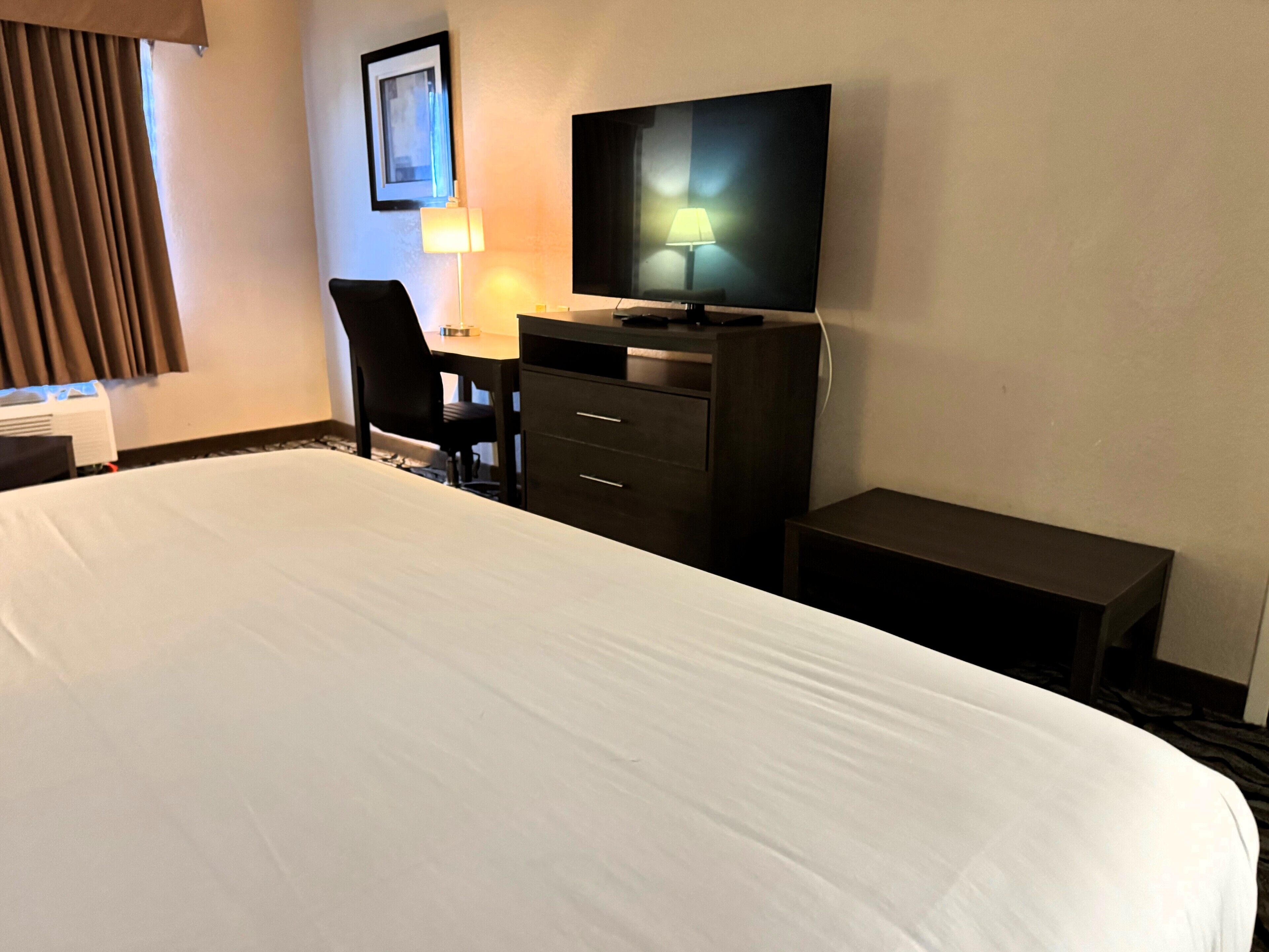 A hotel room with a large bed, a desk with a chair, a television, and a lamp.