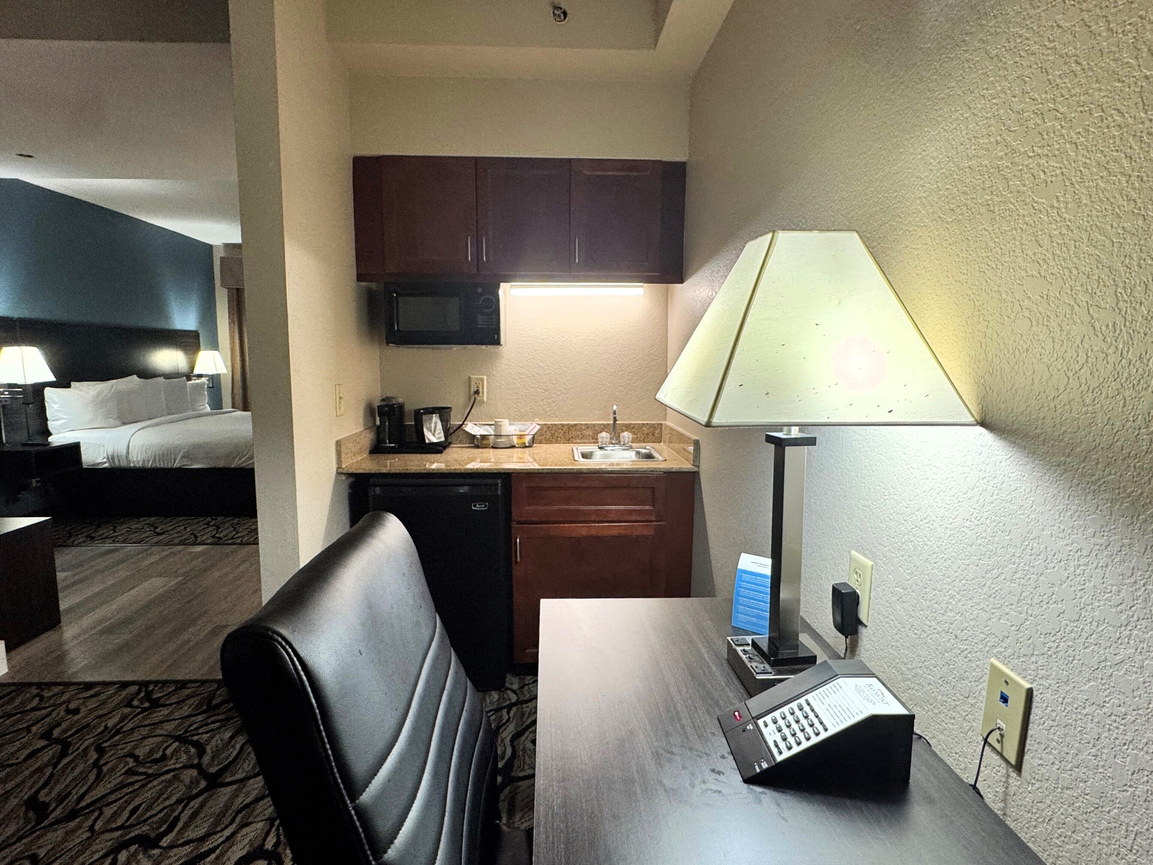 A hotel room with a bed, a desk with a lamp, a microwave, a coffee maker, and a small kitchenette.