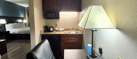 A hotel room with a bed, a desk with a lamp, a microwave, a coffee maker, and a small kitchenette.