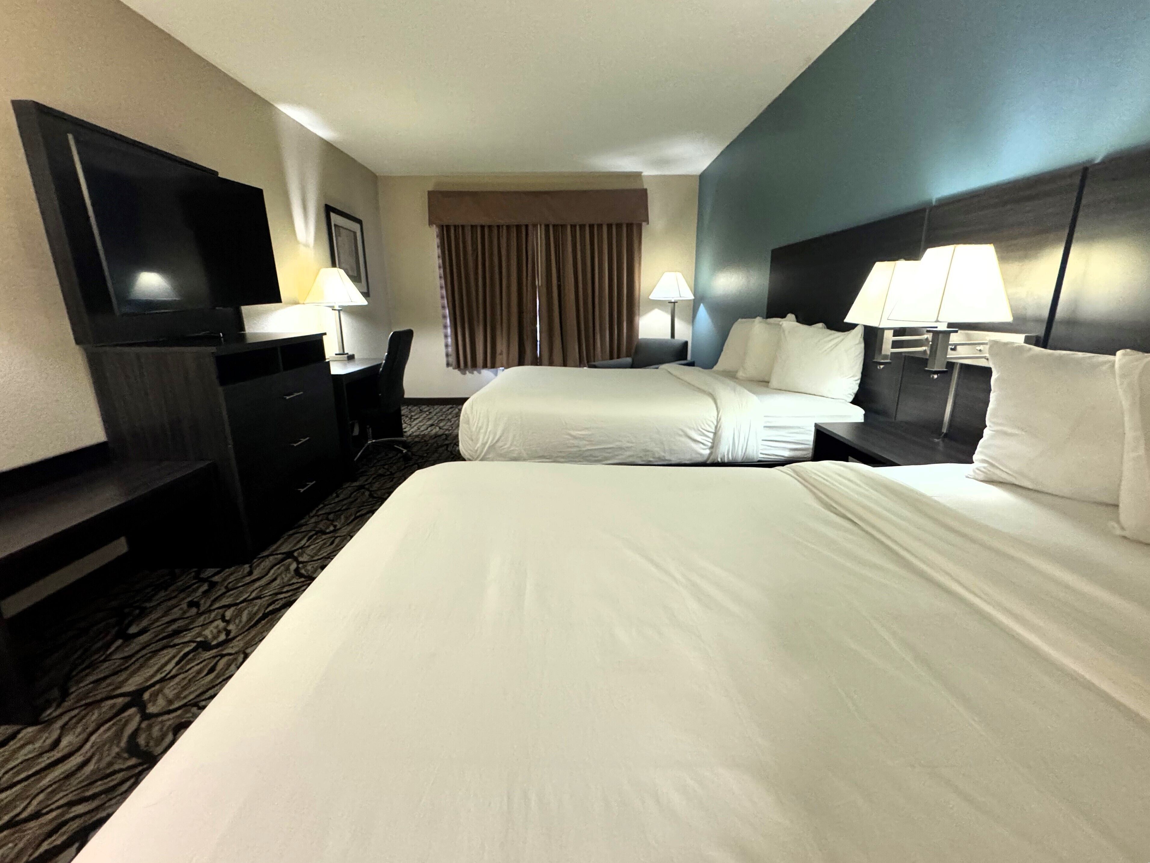 A hotel room with two beds, a desk, a chair, a TV, and lamps.