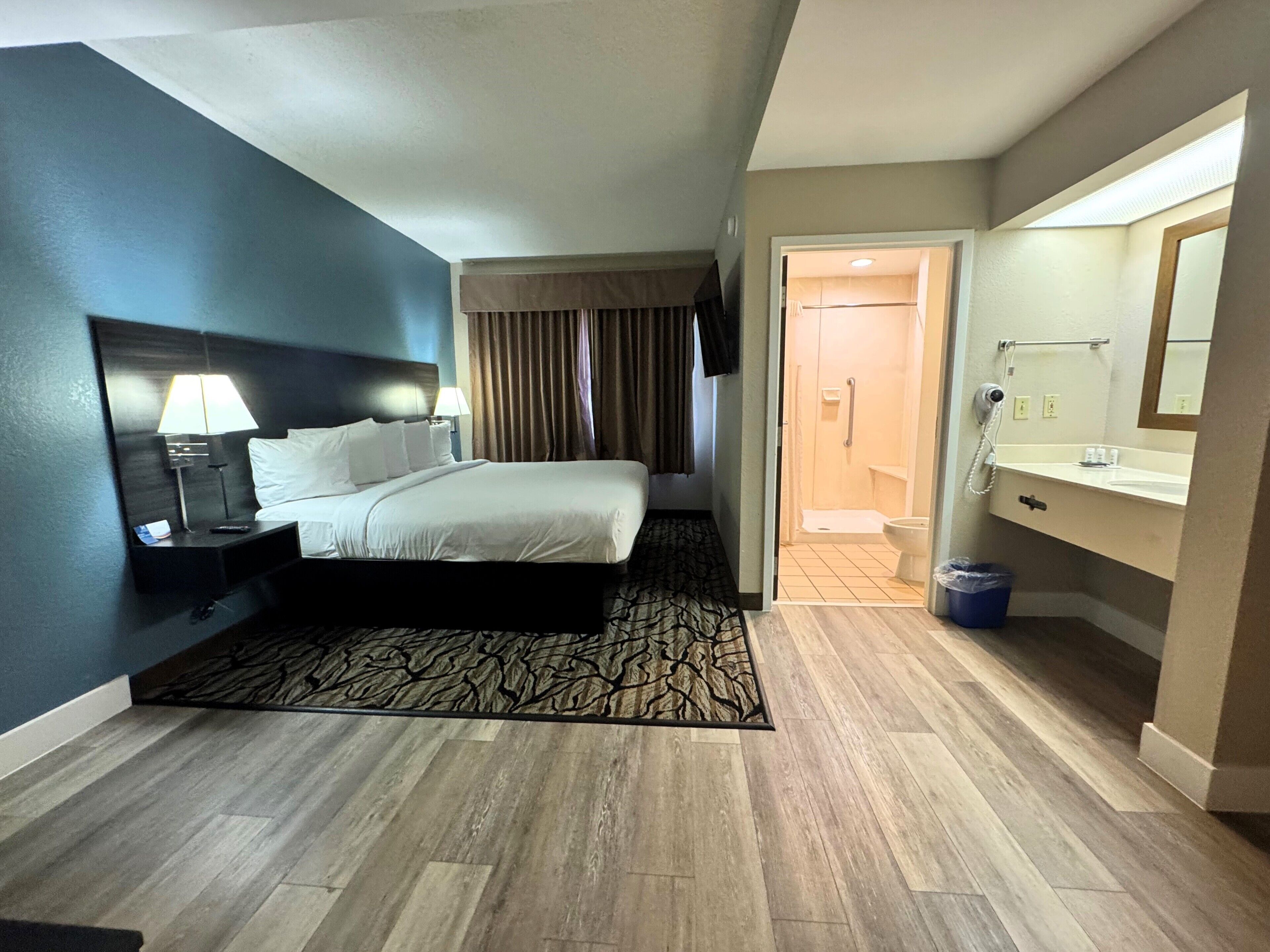 A hotel room with a bed, bedside table with lamp, a bathroom with sink and mirror, and a walk-in closet.