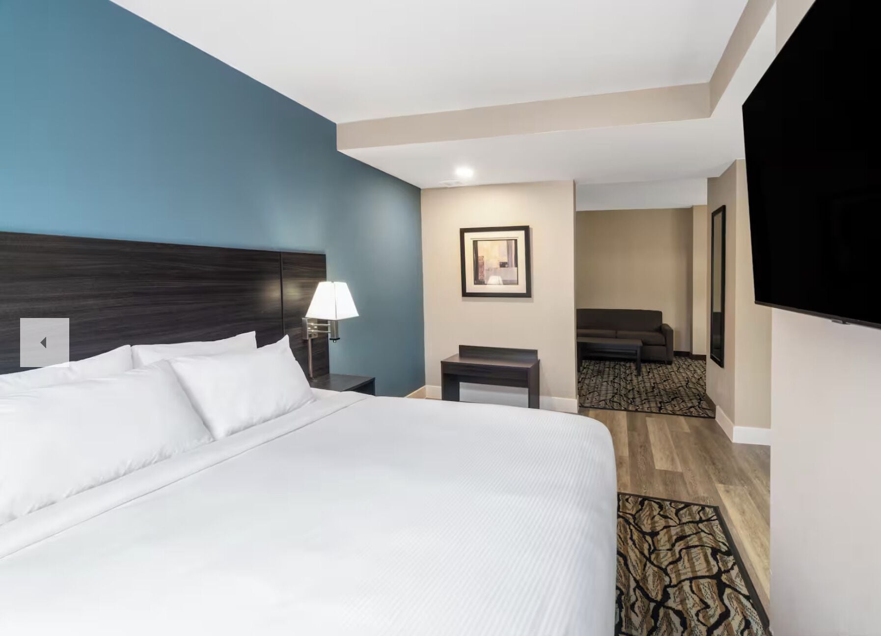 A hotel room with a large bed, a nightstand with a lamp, a small table, a framed picture on the wall, a sofa, and a TV.