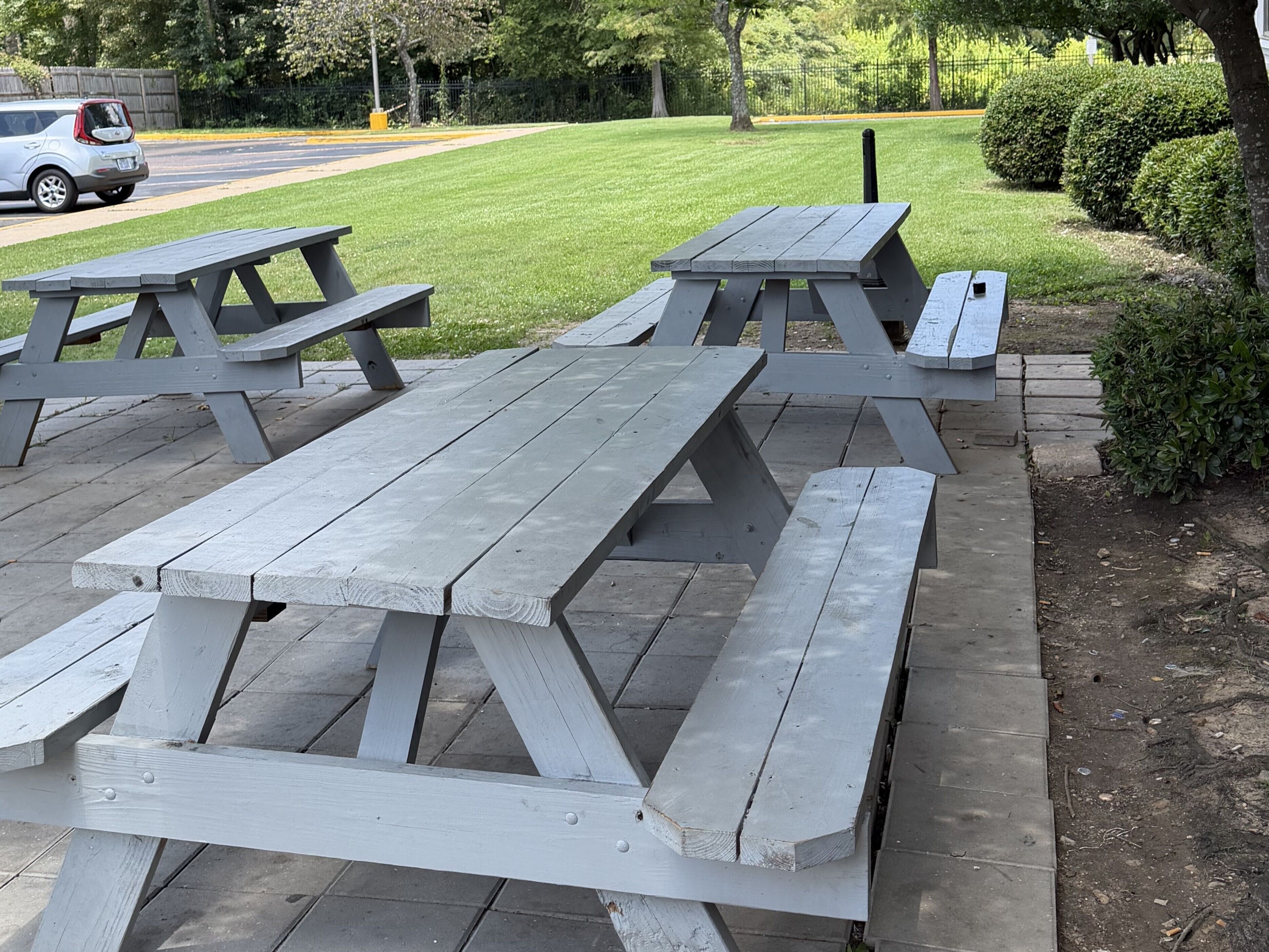 A picnic table set on a paved area with a grassy field and trees in the background.