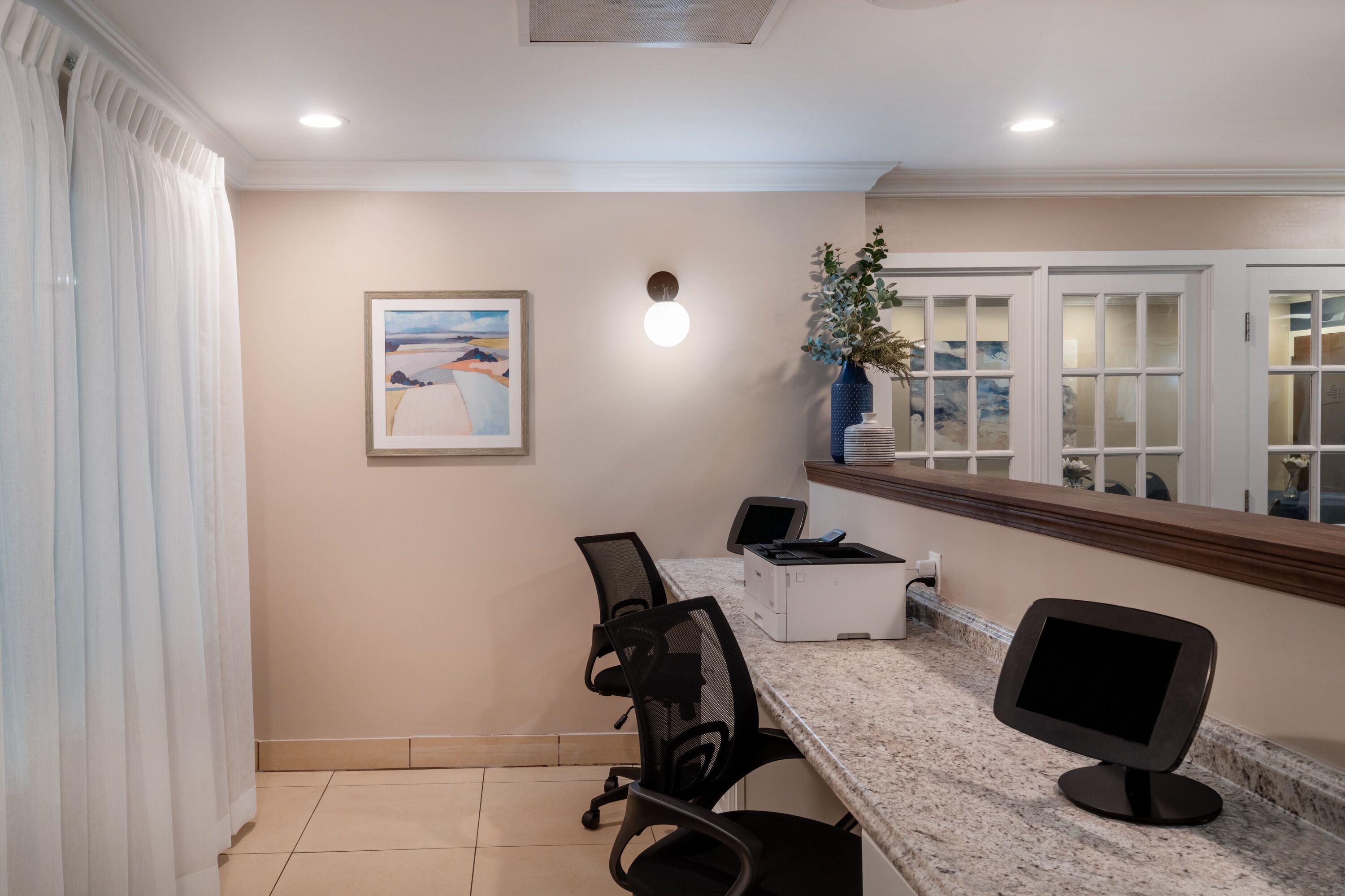A conference room with a marble countertop, two office chairs, a printer, a monitor, a framed painting, and a potted plant.