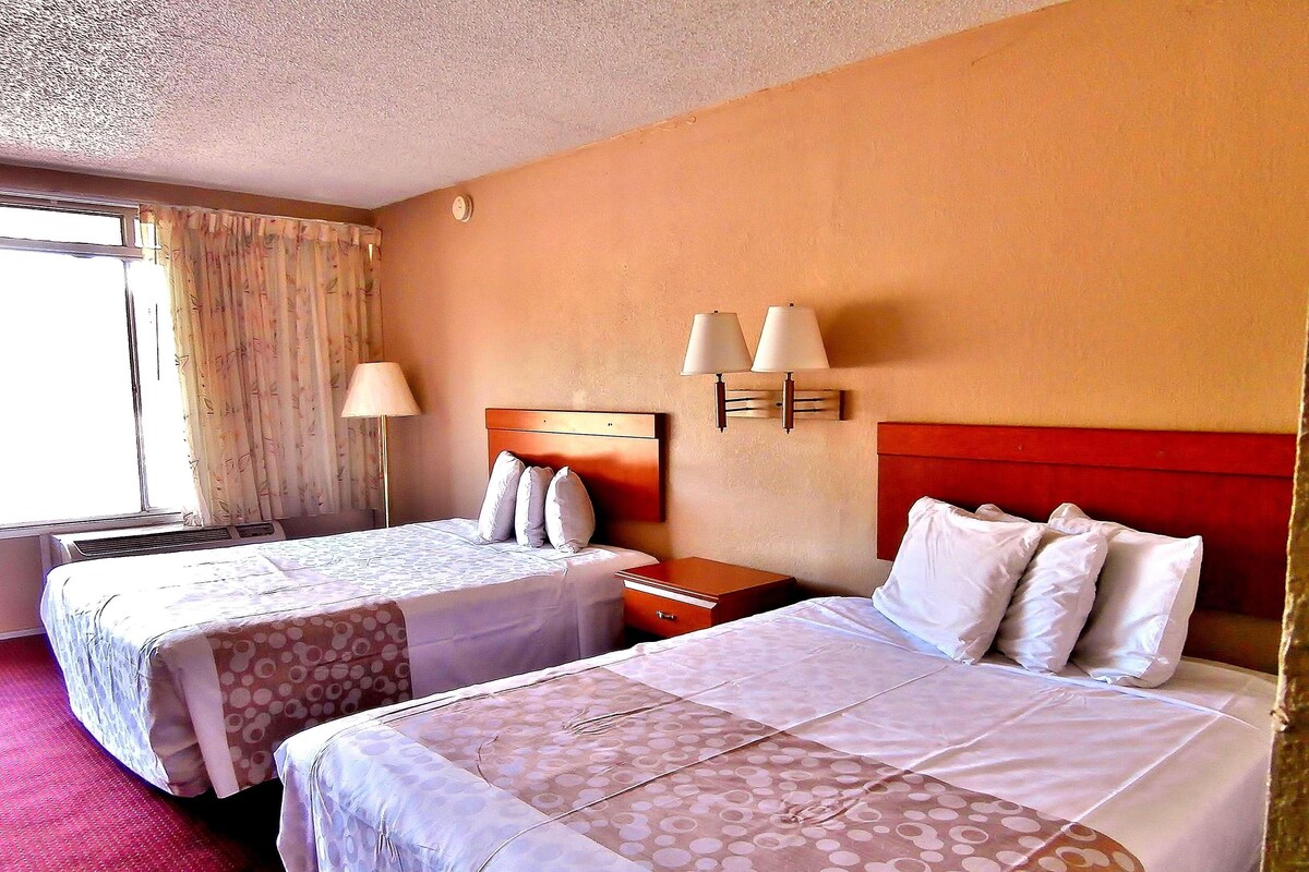A hotel room with two beds, a nightstand, a lamp, and a window with curtains.