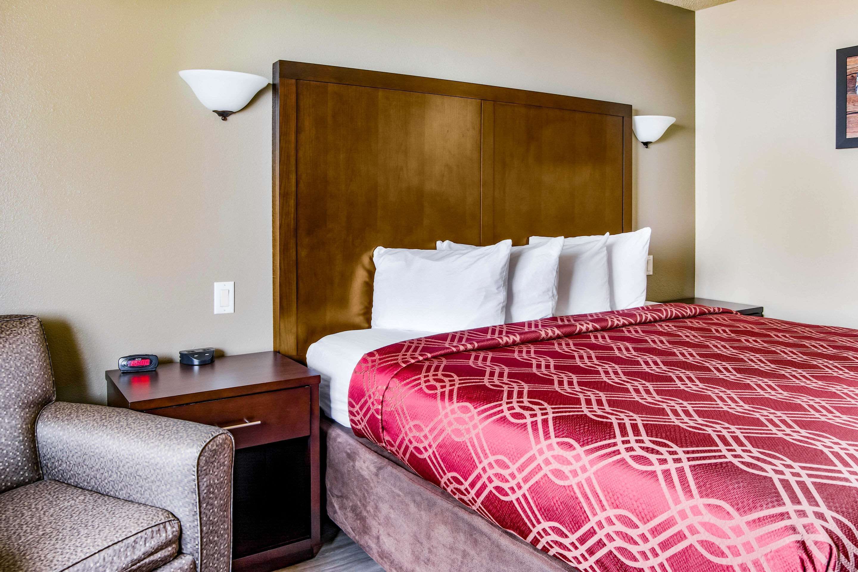 A hotel room with a wooden headboard, a red patterned bedspread, white pillows, a wooden nightstand with a clock, and a gray armchair.