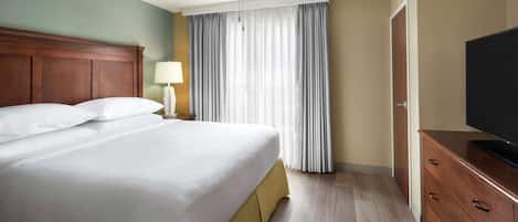Una habitación de hotel con una cama grande, una mesita de noche con lámpara, un televisor sobre un aparador de madera y una ventana con cortinas.