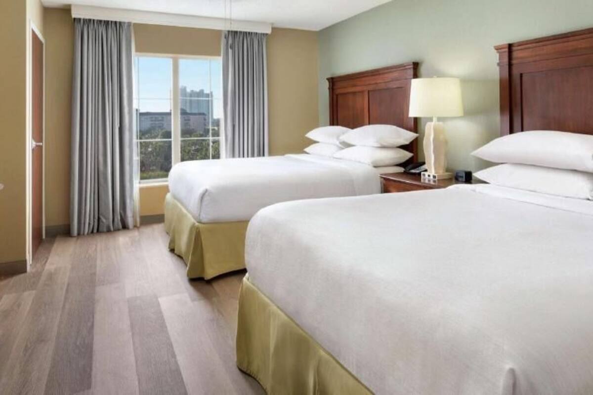 Une chambre d’hôtel avec deux lits, une télévision, une commode, une fenêtre avec des rideaux et une table de chevet avec une lampe.