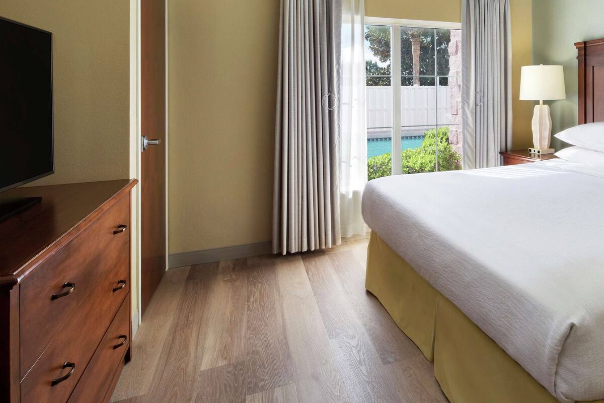 Une chambre d’hôtel avec un lit en bois, une table de chevet avec une lampe, un téléviseur posé sur une commode en bois, et une vue sur une piscine et la verdure par la fenêtre.