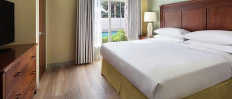 A hotel room with a wooden bed, a nightstand with a lamp, a TV on a wooden dresser, and a view of a pool and greenery through the window.