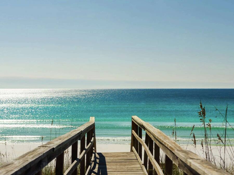 Ein hölzerner Steg führt zu einem Strand mit klarem, blauem Wasser und einem strahlend blauen Himmel.