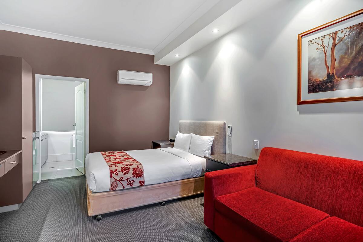 A hotel room with a bed, a red sofa, a painting on the wall, and a door leading to a bathroom.