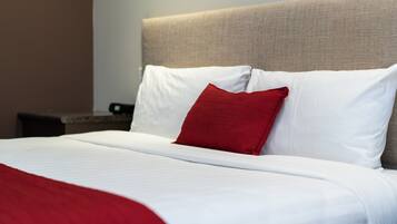 Una cama bien hecha con ropa de cama blanca y roja, un cabecero beige y una mesita de noche con una lámpara.