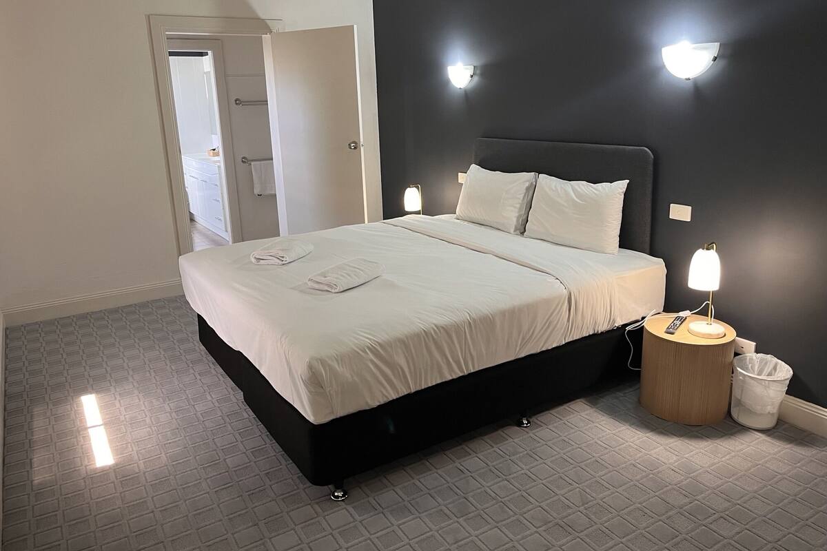 A neatly made bed with white linens and towels, a bedside table with a lamp, and a trash bin.