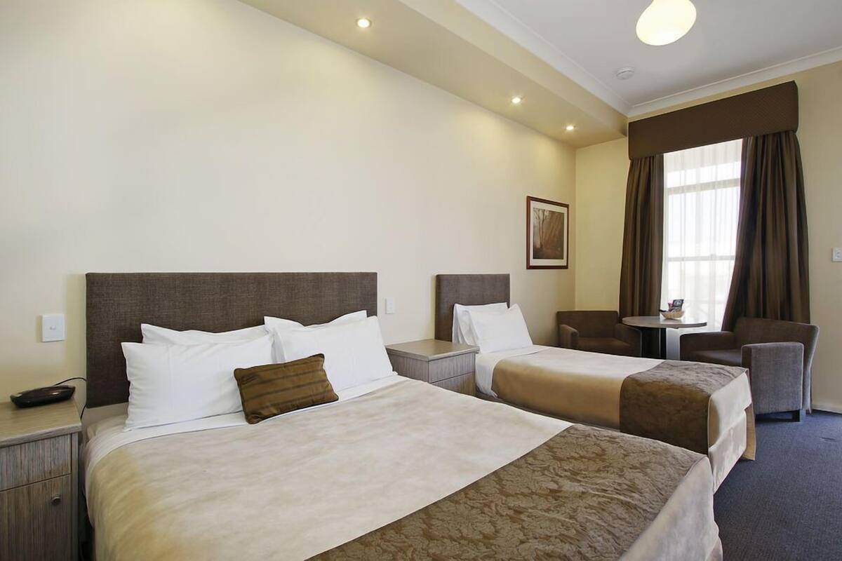 A hotel room with two beds, a small table, a chair, and a window with curtains.
