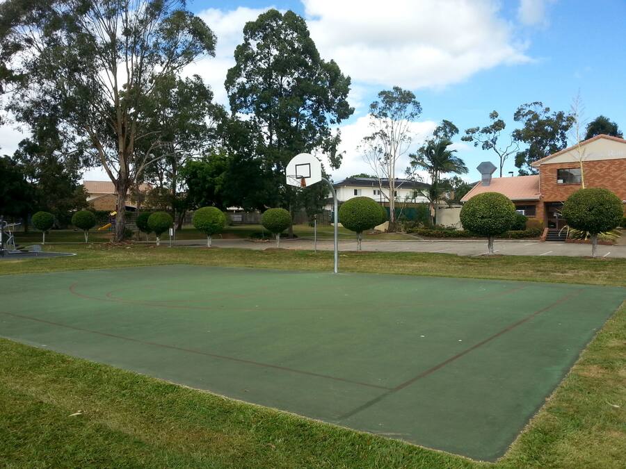 Una cancha de baloncesto al aire libre con superficie verde, rodeada de árboles y edificios residenciales.