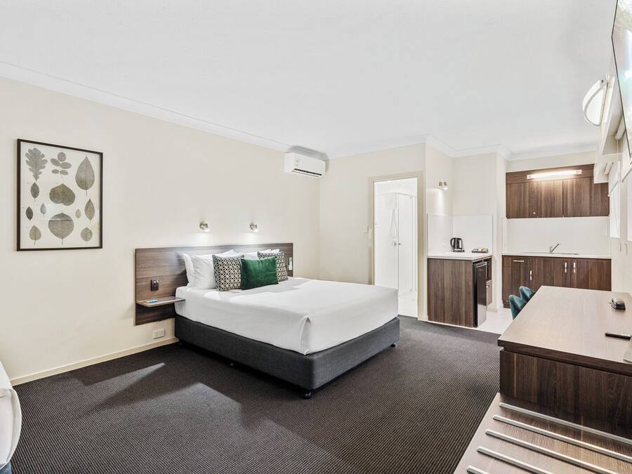 Habitación de hotel con cama, mesita de noche, escritorio pequeño con silla. Cuenta con zona de ducha y kitchenette con lavamanos y cafetera.