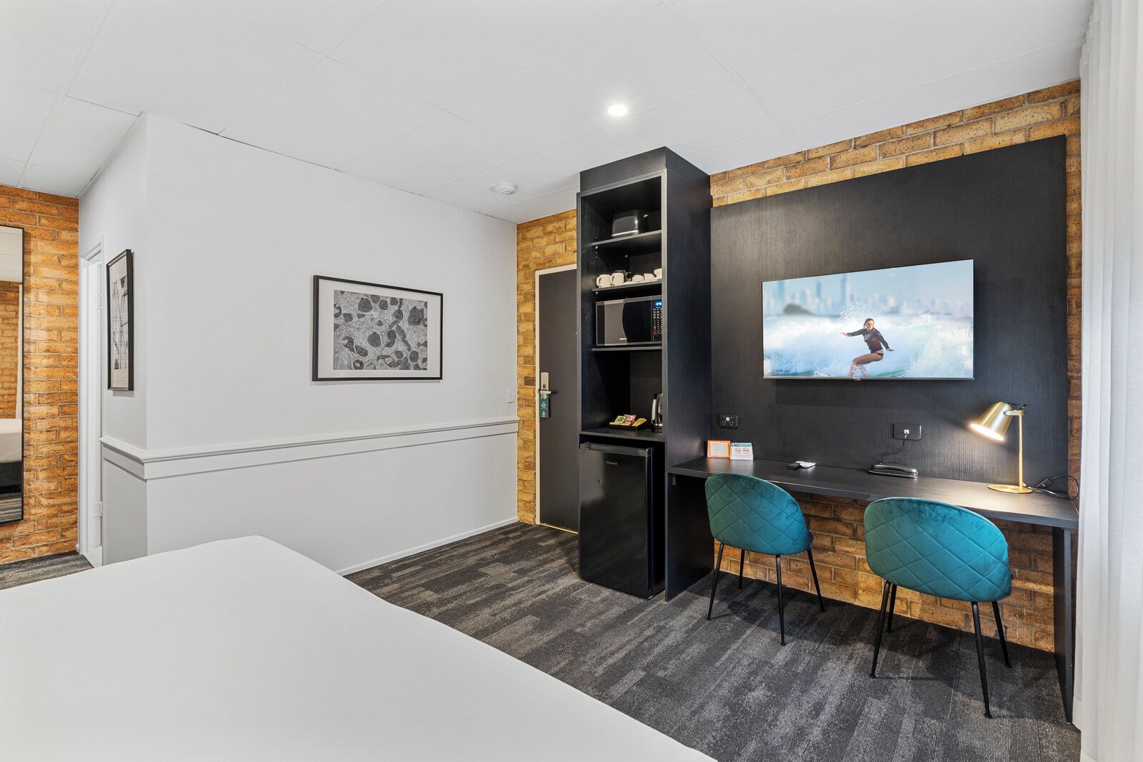 A modern room with a white wall, a black entertainment unit, two teal chairs, a white table, and a mounted TV displaying a surfer.