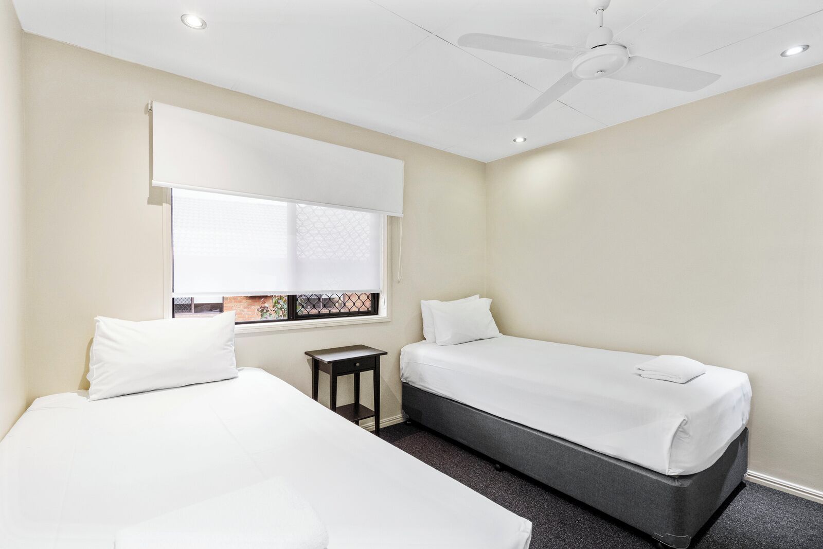 A hotel room with two beds, a ceiling fan, a window with blinds, and a small table.