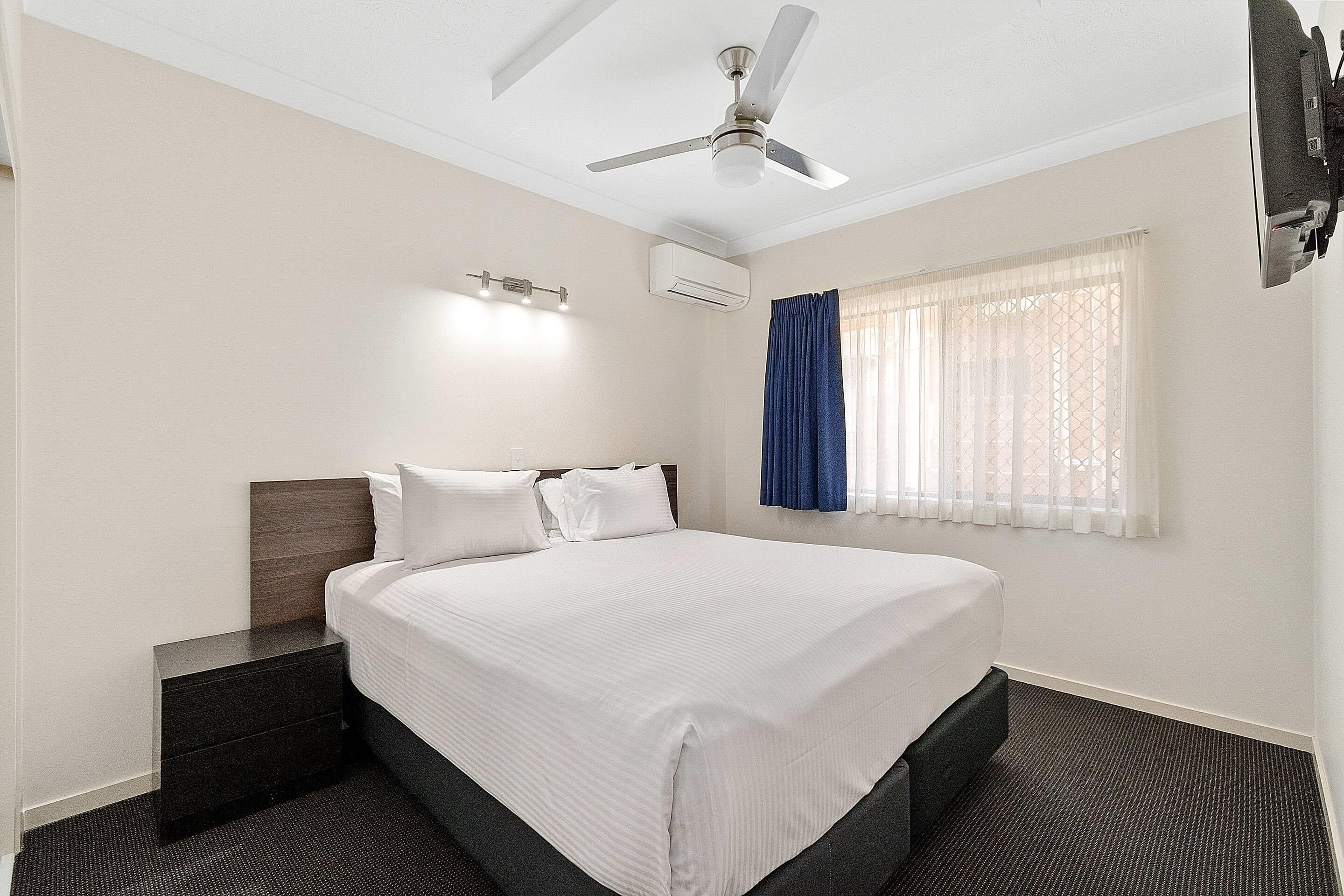 A hotel room with a bed, a nightstand, a ceiling fan, an air conditioning unit, a window with curtains, and a flat-screen TV mounted on the wall.