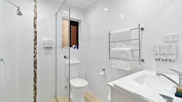 Un baño moderno con ducha envuelta en vidrio, un lavamanos blanco con encimera de mármol y un perchero con varias toallas.