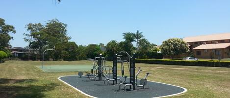 An outdoor gym area with exercise equipment, surrounded by grass and trees, with a building in the background.