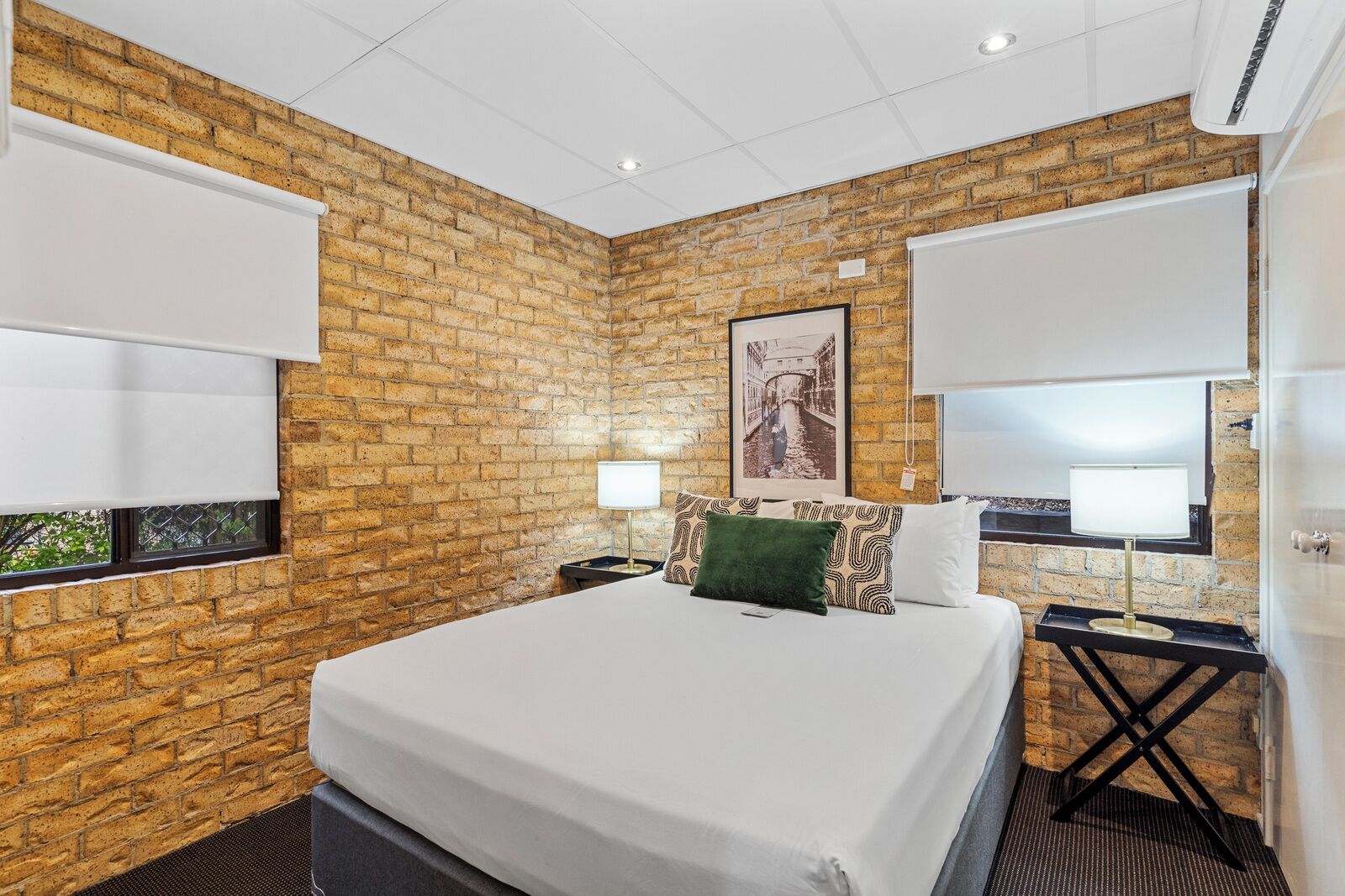 Habitación de hotel con pared de ladrillo, una cama grande, dos mesitas de noche con lámparas y una ventana con persianas.