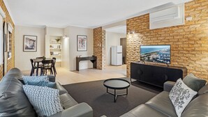 Un salon moderne avec un canapé, une table basse et un téléviseur fixé à un mur en briques. On y trouve également un coin repas avec une table et des chaises, ainsi qu’une cuisine équipée d’un réfrigérateur et d’un four à micro-ondes.