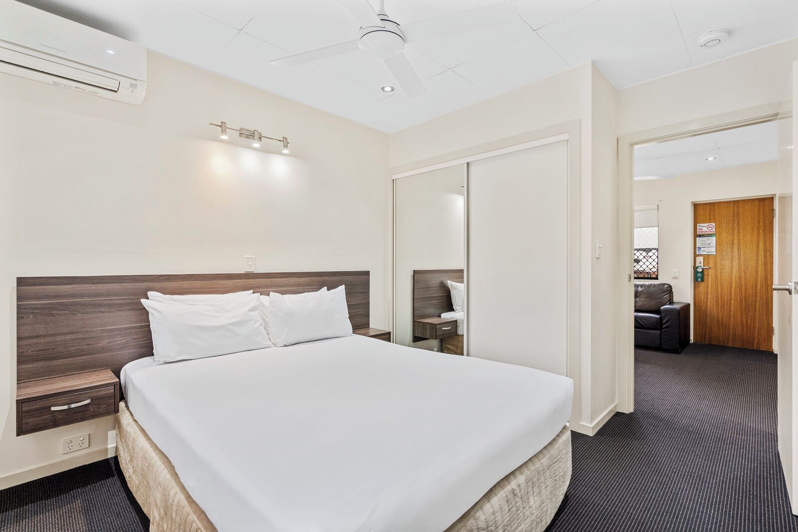 A hotel room with a large bed, a ceiling fan, an air conditioning unit, a bedside table, and a nightstand.