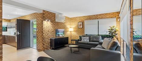 A modern living room with a stone accent wall, a flat-screen TV, a sofa, a coffee table, and a dining area with a table and chairs.