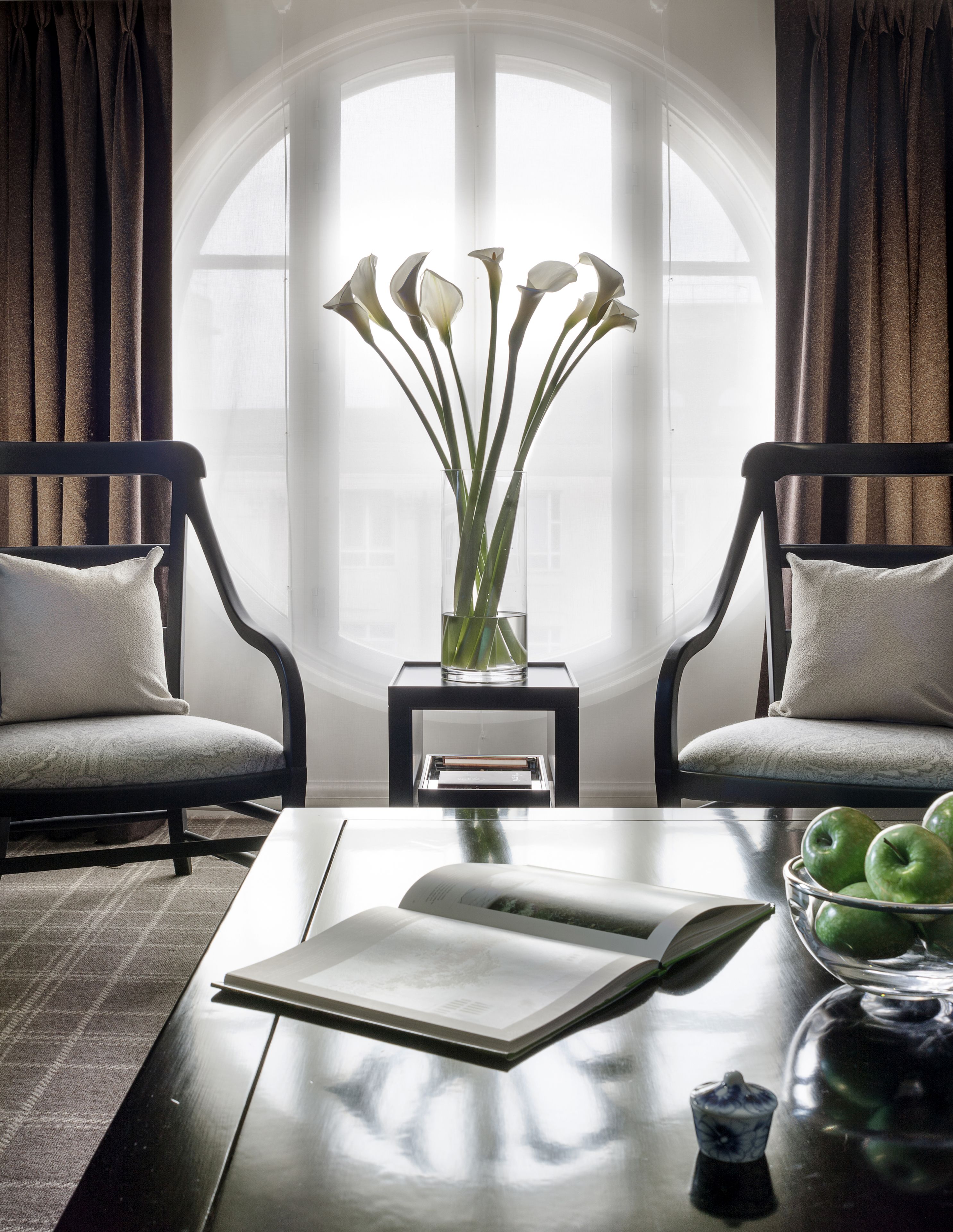 Ein Zimmer mit zwei Sesseln, einem Couchtisch mit einem Buch und einer Schüssel grüner Äpfel, sowie einer Vase mit Blumen.