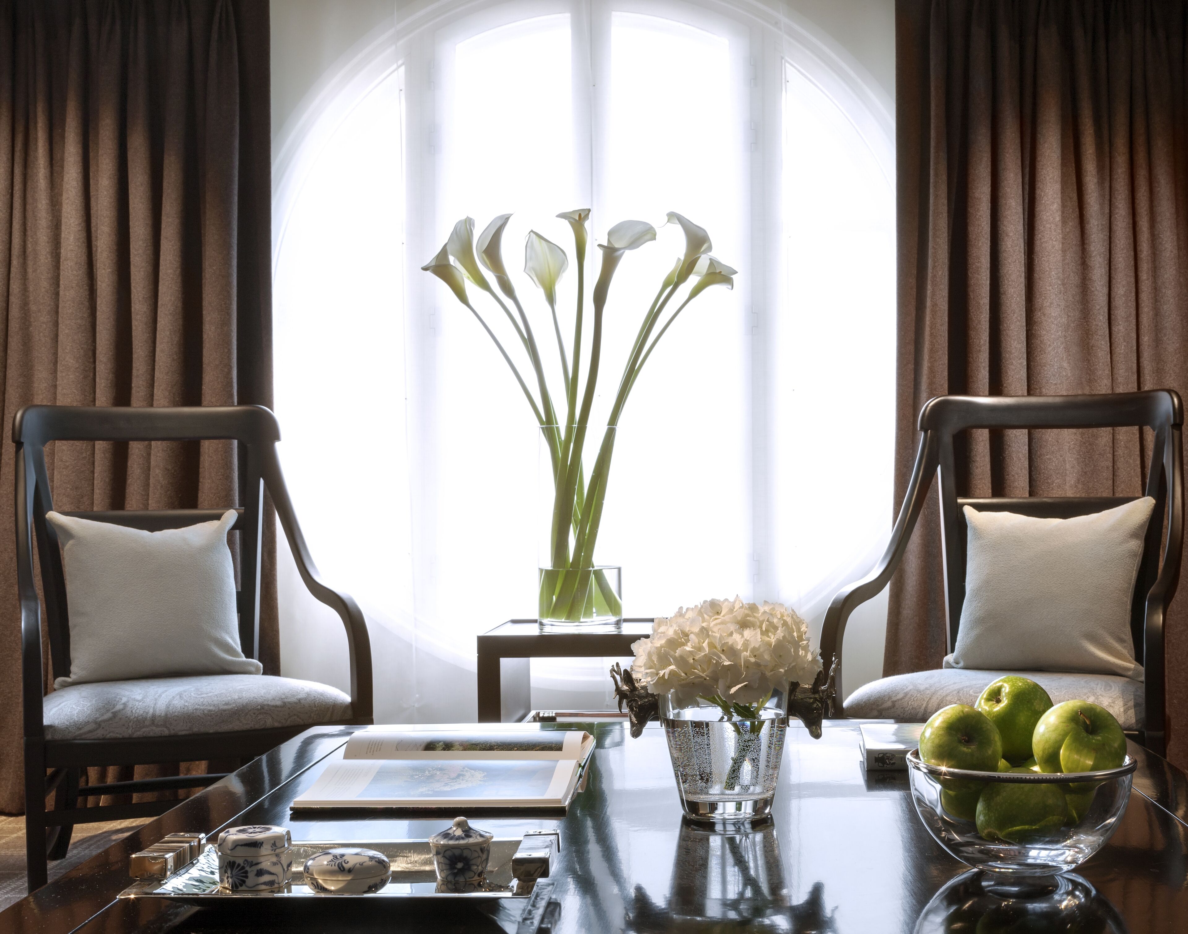 A room with two chairs, a table with a vase of flowers, a bowl of apples, and a book.