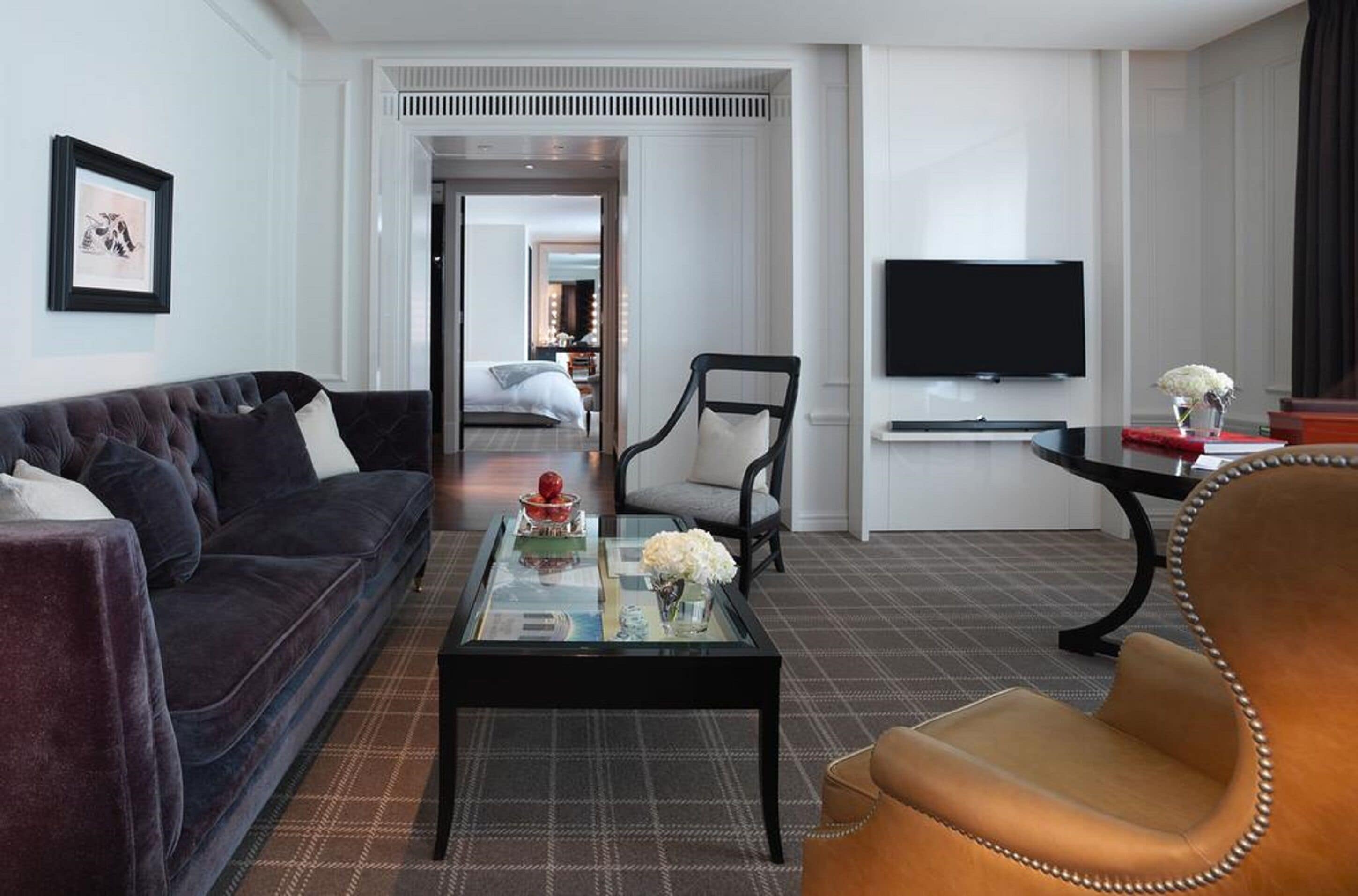 Ett modernt hotellrum med en soffa i mörk färg, ett soffbord i glas med dekorativa föremål, en fåtölj i läder, en platt-TV som är monterad på väggen och en matta med rutmönster.