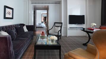 Ett modernt hotellrum med en soffa i mörk färg, ett soffbord i glas med dekorativa föremål, en fåtölj i läder, en platt-TV som är monterad på väggen och en matta med rutmönster.